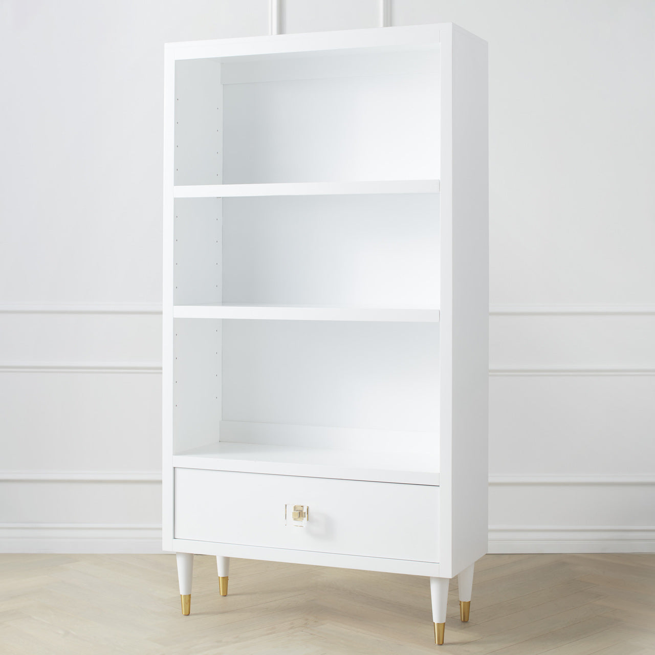 A white Uptown Bookcase with a drawer and three shelves, featuring brass or chrome lucite pulls on a white background.